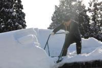 男性が屋根の上で、大きな羽子板のような道具を使って雪下ろしをしている写真