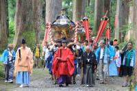 宮司を先頭に神輿を担いだ人や青い法被を着た人々が列をなして木々の間の道を歩いている写真