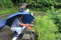 傘を差している男性が、道路脇の石でできた台の上にお供え物を置いている写真