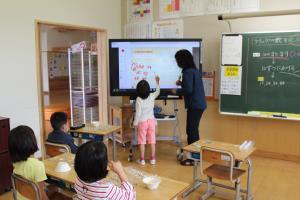 電子黒板が置かれた教室の前方で女子児童が電子黒板の前に立って発表しており、その様子を先生と他の児童が見守っている授業風景の写真