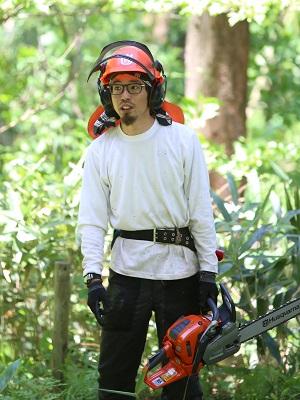 緑が生い茂っている背景の中赤いヘルメットを被り、チェンソーを持っている伊藤 章吾さんの写真