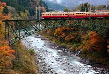 紅葉で綺麗に色づいている山の下に川が流れており、鉄橋の上を列車が走っている写真