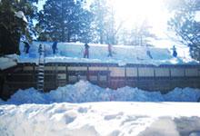 家の前や屋根に沢山の雪が降り積もり、屋根に梯子をかけて7名の人が屋根に上って雪下ろしをしている写真