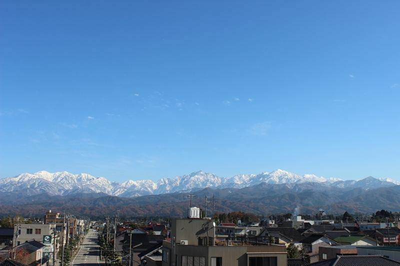 遠くに雪が積もっている立山連峰が見え、街並みが広がっているみらいぶからの眺めの写真