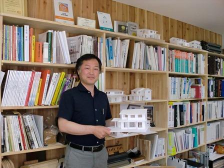 沢山の本が並んでいる本棚の前に2階建ての家の模型を持って写っている前川建築さんの写真