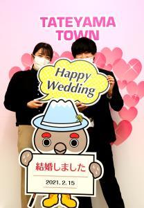 ハート模様のお祝いボードの前で 「Happy Wedding」と書かれたをフォトプロップス持った男女2人が「結婚しました。2021年2月15日」と書かれたボートを持ったマスコットキャラクター「らいじぃ」 の後ろに並んで写っている写真
