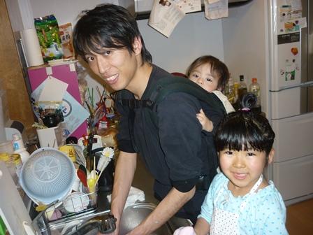エプロンを付けた女の子と赤ちゃんをおんぶしているお父さんがキッチンのシンクのところで作業をしている写真