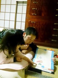 布団の上に腹ばいで寝転がって女の子と一緒に絵本を読んでいるお父さんの写真