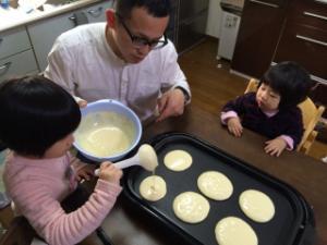ホットプレートにホットケーキの生地をお玉で注いでいる女の子を見守っているお父さんの写真
