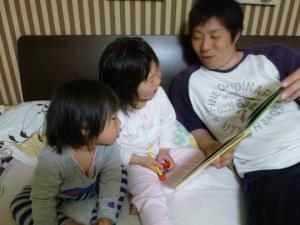 布団の上で子供達2人に絵本を読んでいるお父さんの写真