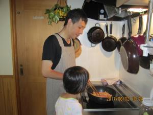 女の子が見守る中、エプロンを付けてキッチンで料理をしているお父さんの写真