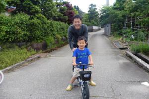 自転車に乗っている男の子の後ろに笑顔で立っているお父さんの写真