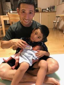 男の子を膝の上に乗せ、その上に赤ちゃんを乗せてミルクを飲ませているお父さんの写真