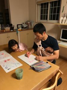 ベビースリングを使って赤ちゃんを抱っこしながら女の子と一緒にテーブルに座り絵を描いているお父さんの写真