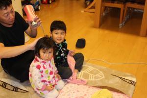 床に座り男の子と女の子の髪をドライヤーで乾かしているお父さんの写真