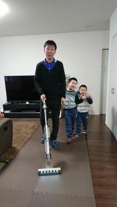 お父さんが掃除機をかけている後ろに男の子2人がいる写真