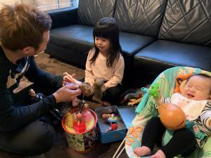 赤ちゃんがバウンサーに乗っている横で女の子とお父さんが人形遊びをしている写真