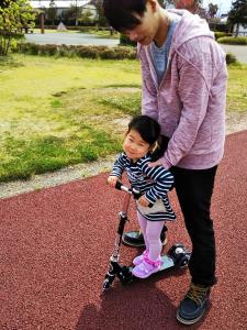 キックボードに乗る女の子を後ろからサポートしているお父さんの写真