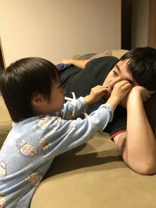 寝転がっているお父さんの顔を触って遊んでいる男の子の写真