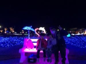 イルミネーションの会場で子供二人とベンチに座りピースサインをしているお父さんの写真