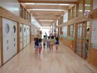 奥へと長く続いている木製で明るい廊下を歩いている園児たちの後ろ姿の写真